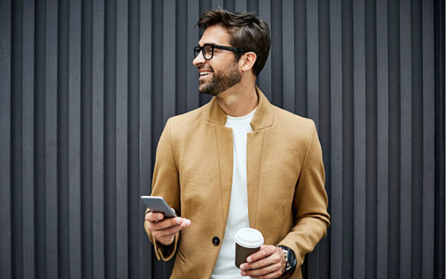 Mann schaut lächelnd nach rechts und hält Smartphone und Kaffee to Go Becher in der Hand
